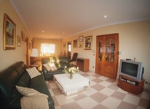 Een woonkamer met een zwart-wit geruite vloer, een groene bank, een salontafel, een televisie en lijstjes met foto’s aan de muren.