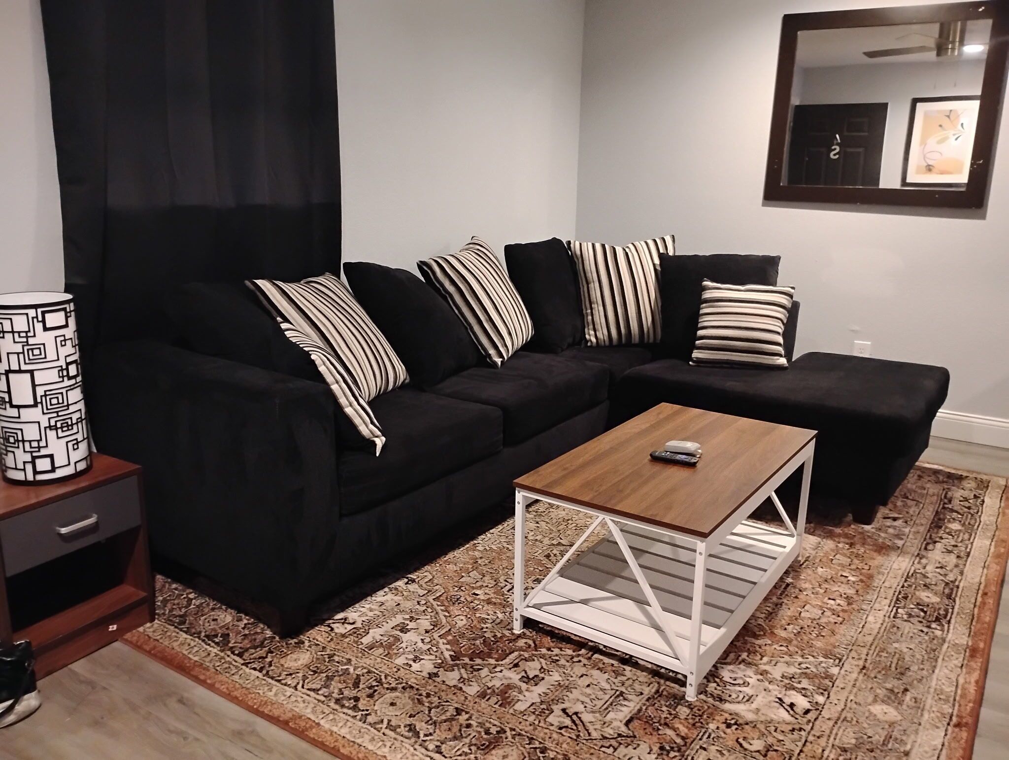 Un salon moderne avec un canapé d’angle noir, des coussins à rayures, une table basse en bois et un tapis à motifs.