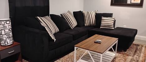 Un salon moderne avec un canapé d’angle noir, des coussins à rayures, une table basse en bois et un tapis à motifs.
