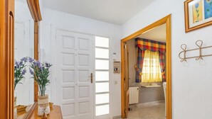 A hallway with a white door, a mirror, a vase with flowers, and a view into a bathroom with a radiator and a towel rack. - Family Oasis in Comarruga: Pool and Relaxation (El Vendrell)