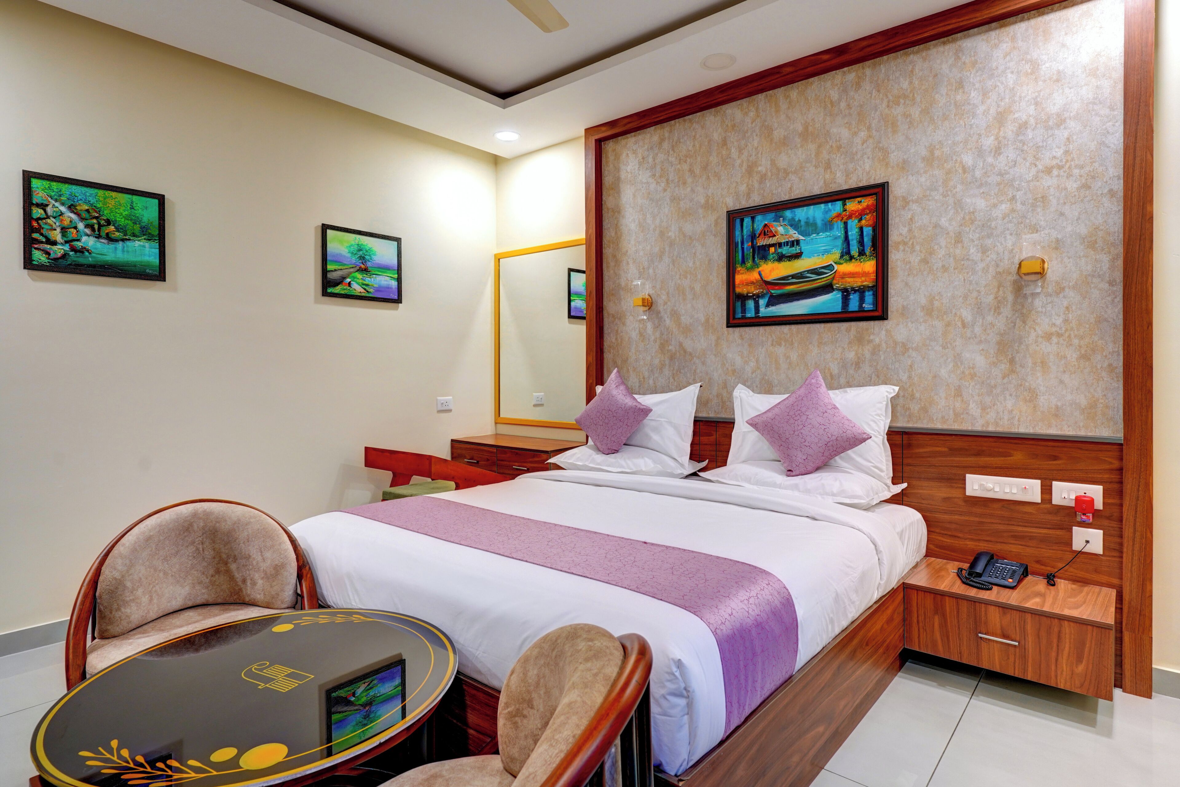 Habitación de hotel con cama, mesitas de noche, una silla, una pequeña mesa y obras de arte en las paredes.