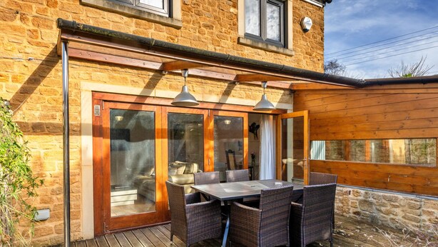 A stone building with a wooden deck, a glass door entrance, wicker furniture, and a window with a shutter. - Hollies Cottage, Oak (3 bed) (Bower Hinton)