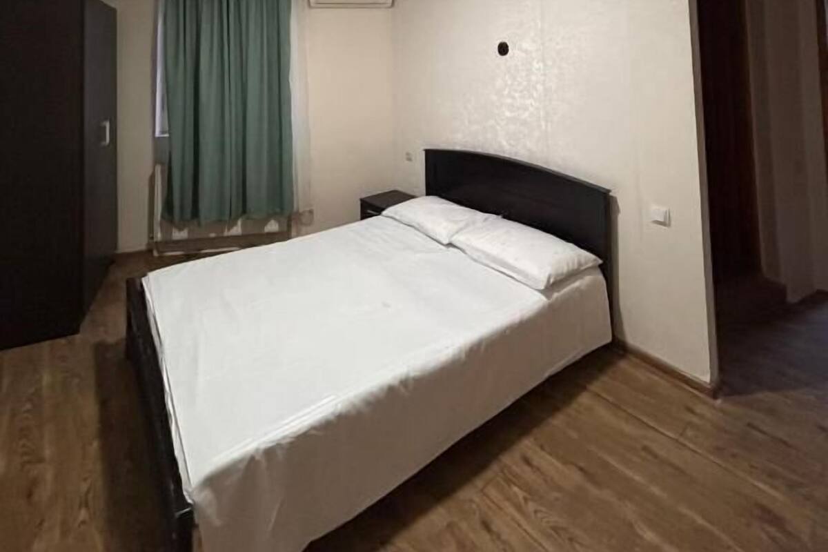 A bedroom with a bed, wooden flooring, a window with curtains, an air conditioning unit, and a door.