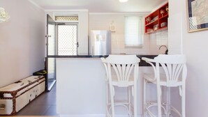 A compact kitchen with a central island, white bar stools, a refrigerator, and a red wine rack.