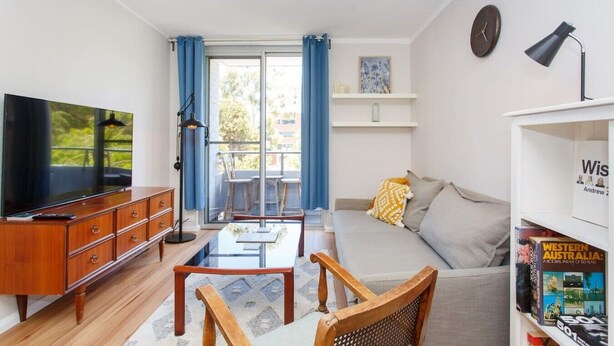 A modern living room with a flat-screen TV, wooden furniture, a bookshelf, and a balcony with blue curtains.