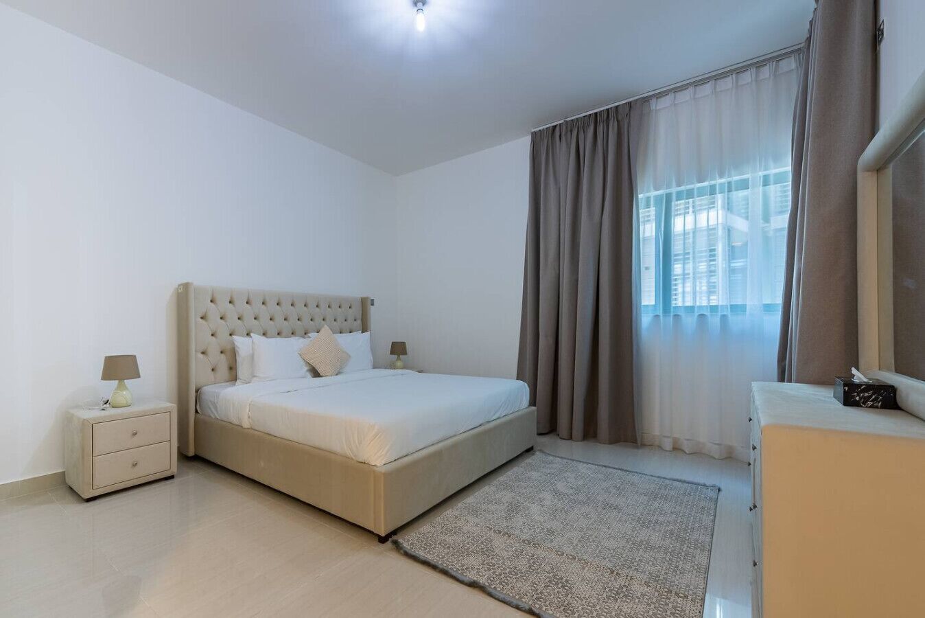 Une chambre à coucher comprenant un lit, des tables de chevet, une commode, une fenêtre avec des rideaux et une vue sur les bâtiments environnants.