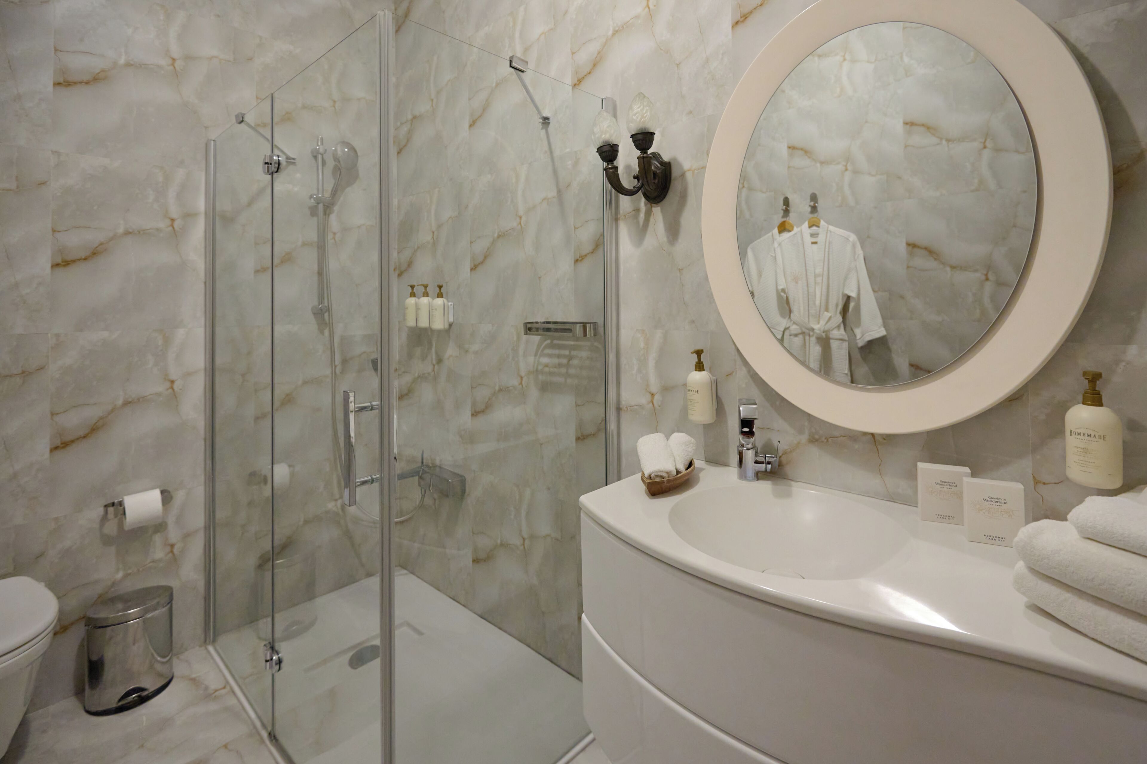 A bathroom with a glass shower enclosure, a marble wall, a round mirror, a sink with toiletries, and a toilet.