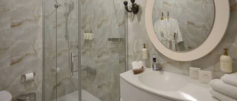 A bathroom with a glass shower enclosure, a marble wall, a round mirror, a sink with toiletries, and a toilet.