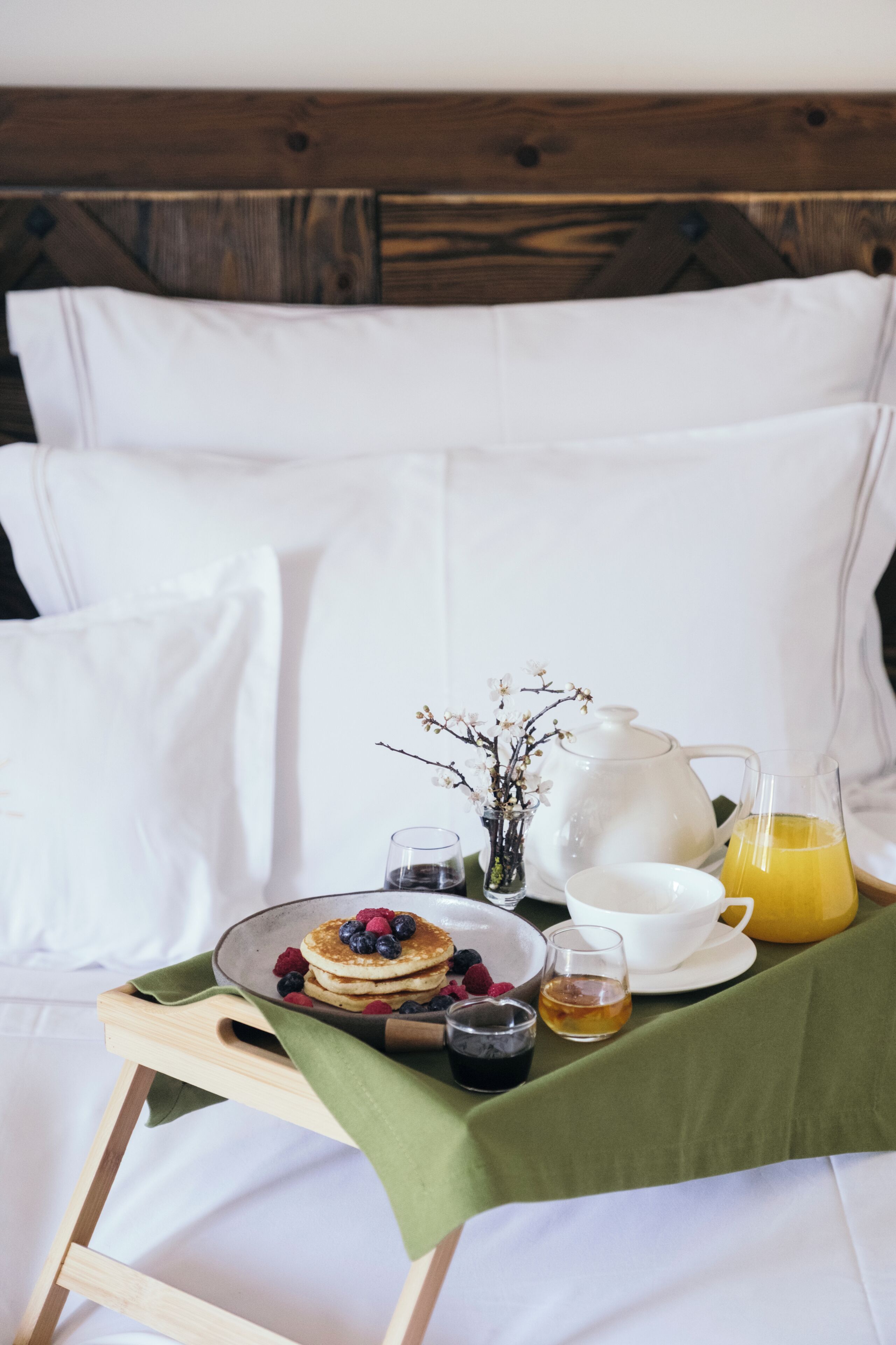 A breakfast setup on a bed with a tray of pancakes, berries, a teapot, cups, and a glass of orange juice.