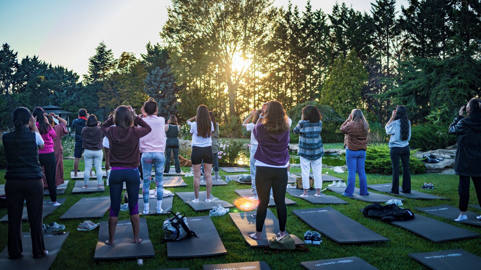 A group of people on yoga mats outdoors with trees and a sun setting in the background.