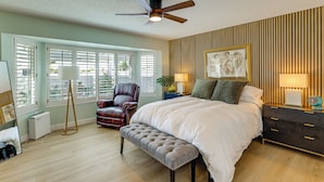 Quarto com cama, ventilador de teto, um banco, uma cadeira, mesa de cabeceira com luminárias e uma obra de arte na parede.