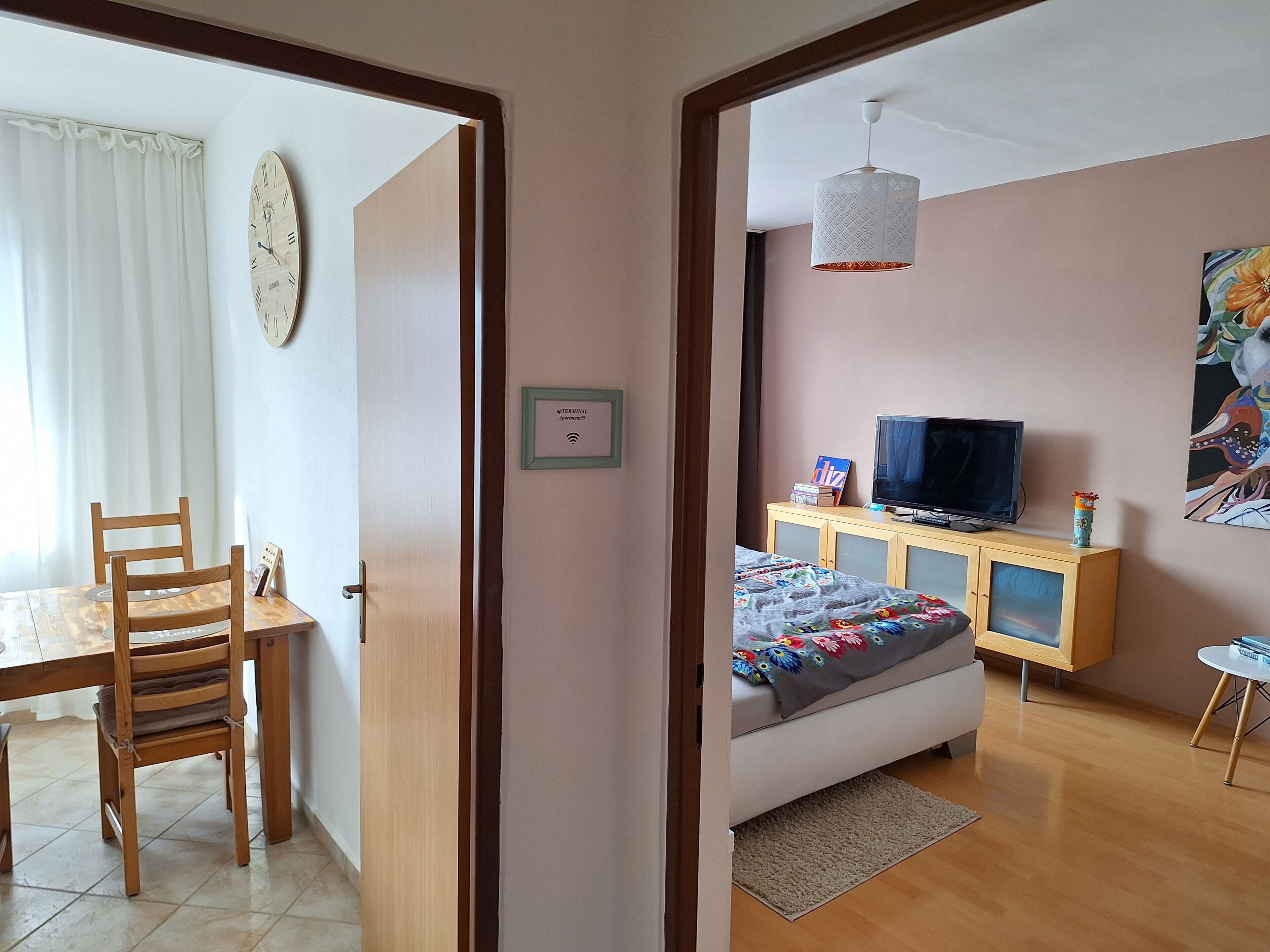 Ein Zimmer mit einem Bett, einem Fernseher, einem Schrank, einem Gemälde und einer Tür zu einem anderen Raum.