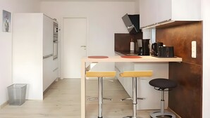 Et lille køkken med træbordplade, to barstole, en kaffemaskine og en skraldespand.