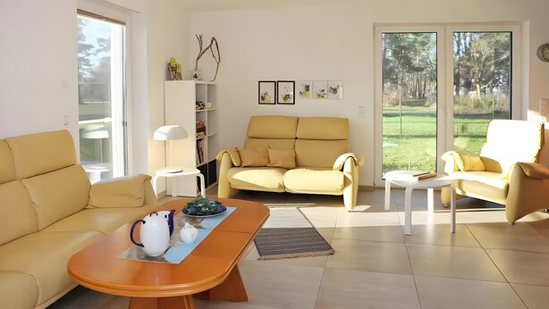 A living room with a sofa, a coffee table, a bookshelf, and a view of a garden through a glass door. - Holiday Home 'Waldwiese' for 8 Guests (110 m²) in Fuhlendorf OT Bodstedt (Boddenküste McPomm) (Fuhlendorf)