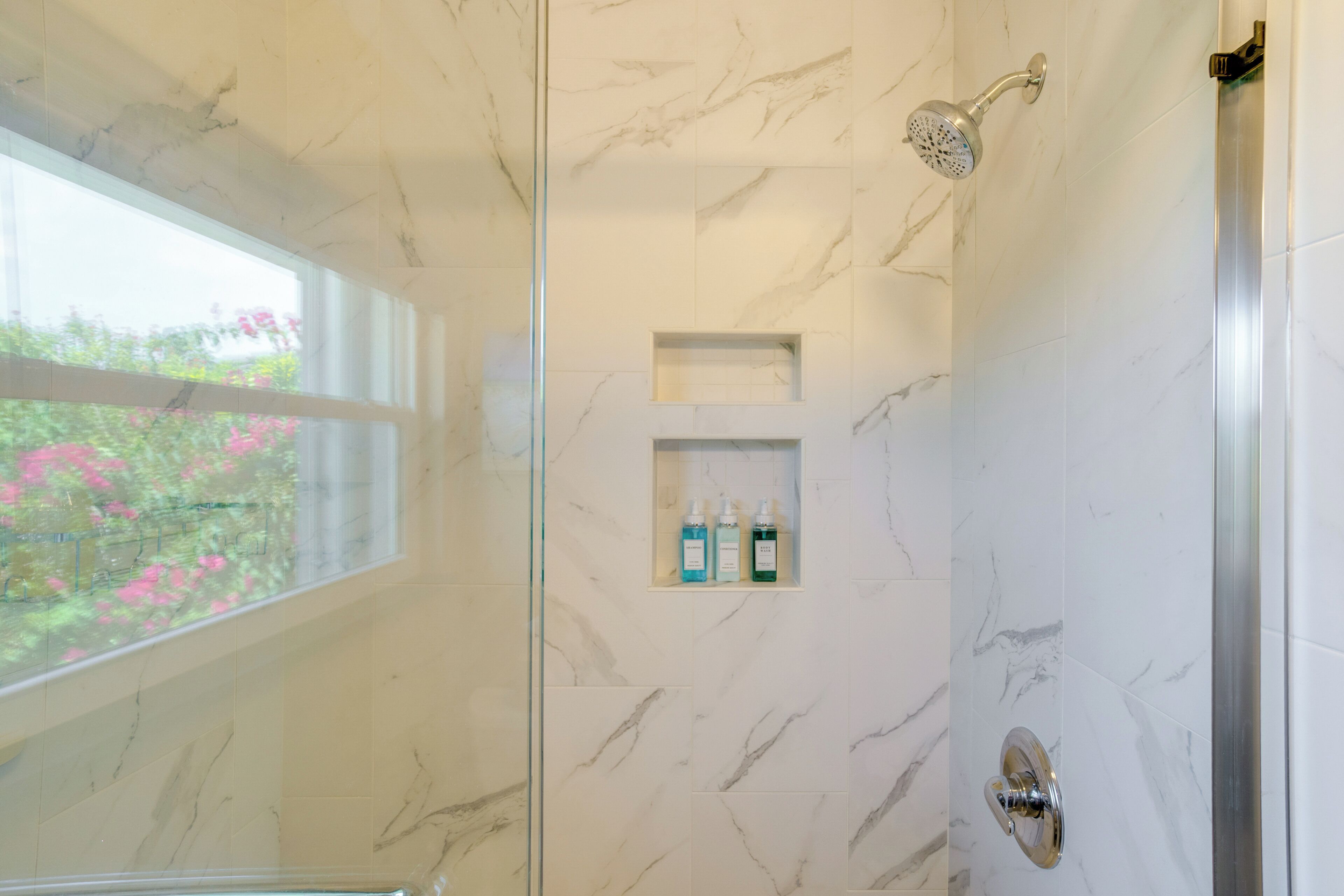 A bathroom with marble walls, a shower with a handheld showerhead, and a window with a view of pink flowers.