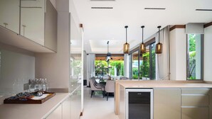 A modern kitchen with a built-in refrigerator, a dining area with a table and chairs, and large windows offering a view of greenery. - Banyan Tree Grand Residences, Four-bedroom Pool Villa (Choeng Thale)