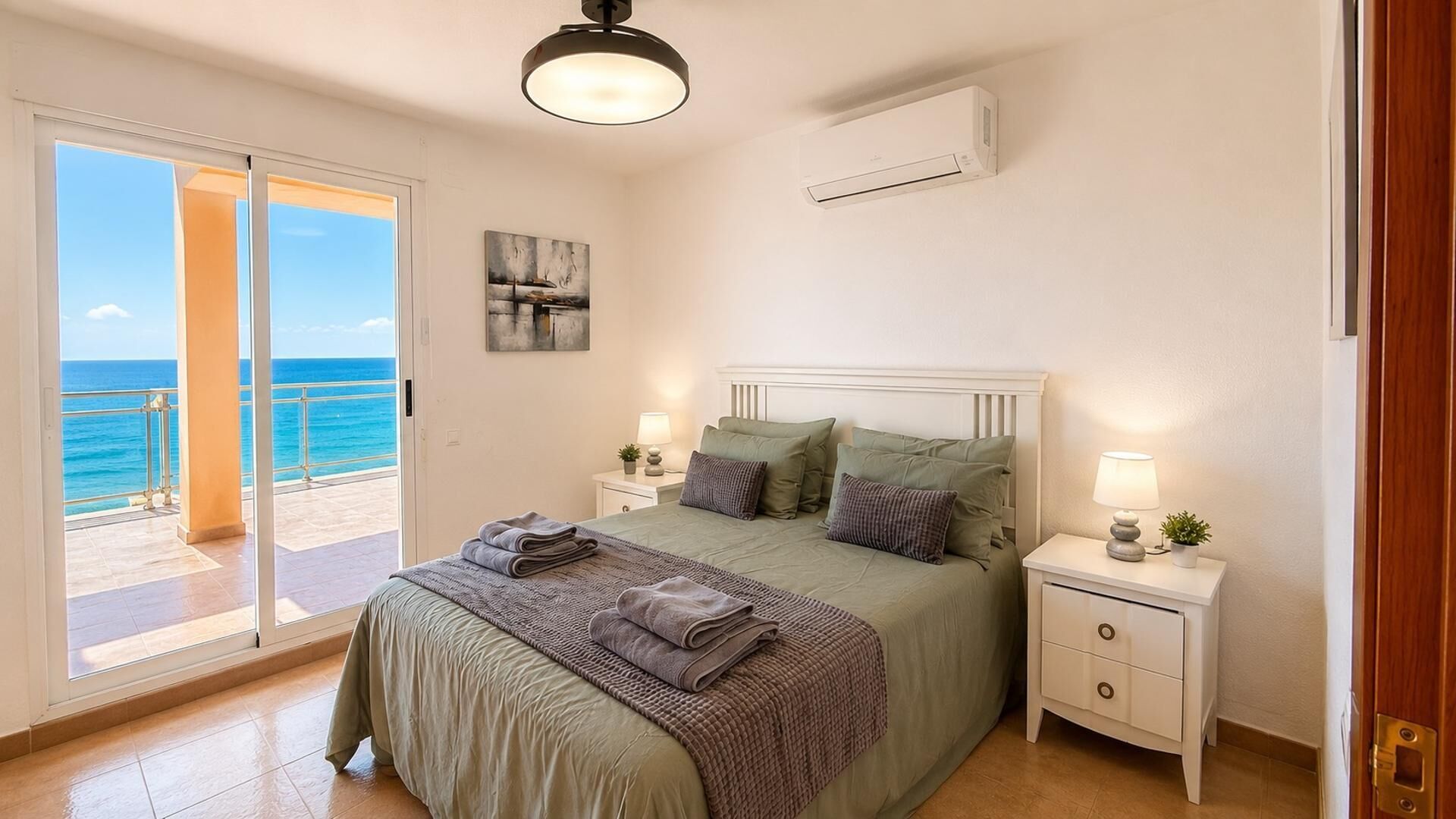 A bedroom with a bed, nightstands, a lamp, a painting, and a view of the sea.