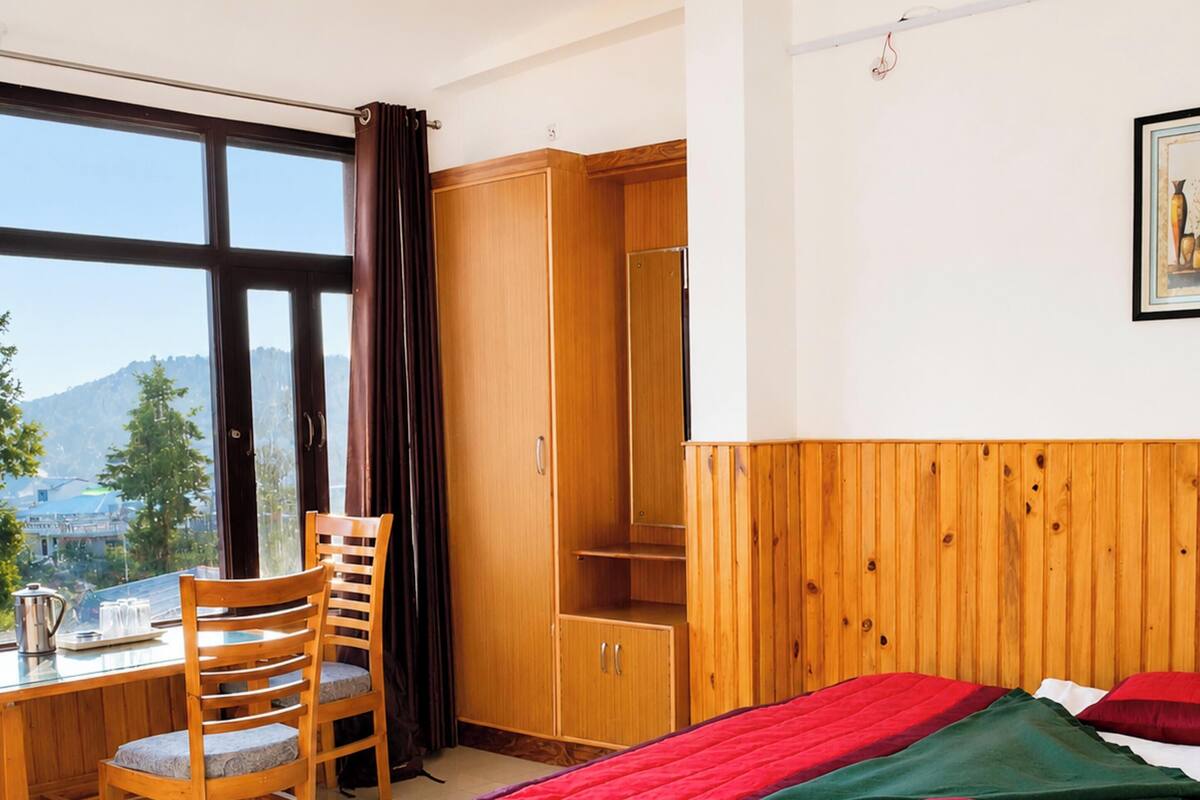 A room with a bed, a desk, a chair, a wardrobe, and a window with a view of a mountain.