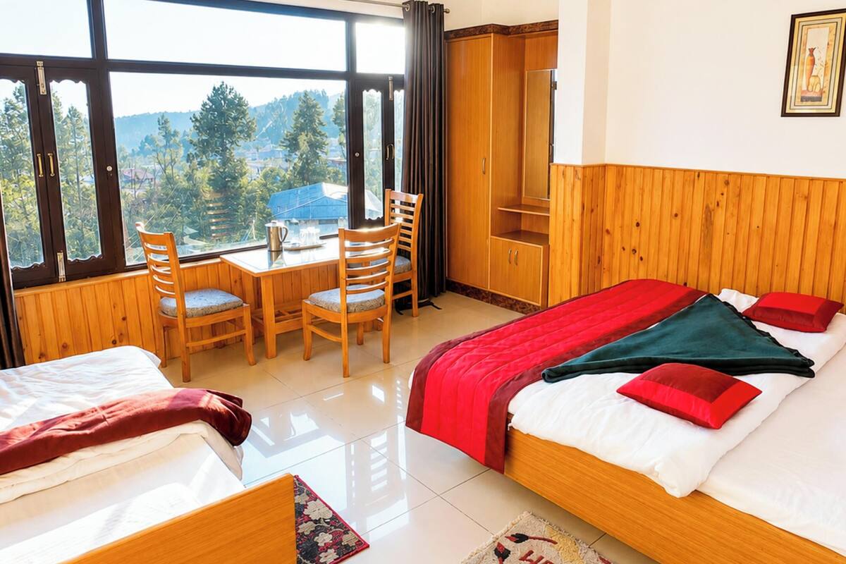 A hotel room with a bed, a desk, two chairs, a wardrobe, and a large window offering a view of trees and buildings.