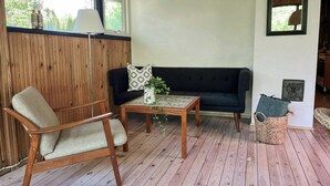 A room with a wooden floor, a black sofa, a wooden chair, a small table with a vase, and a window with a view of trees.