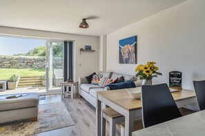 A modern living room with a sofa, a dining table, and a view of a garden through a glass door. - Buttercup Cottage (Wadebridge)