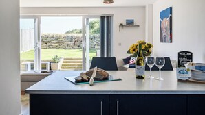 Una cocina moderna con una isla central, un comedor con mesa y sillas, y vistas a una pared de piedra y un jardín a través de puertas correderas amplias.