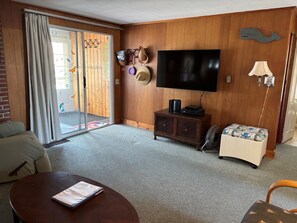 A living room with a sofa, a coffee table, a TV on a wooden wall, a small cabinet, a fan, and a sliding glass door leading outside. - Stay at "Seas the Day" in Ferry Beach, Saco, ME—an inviting 4-bedroom cottage near Camp Ellis (Saco)