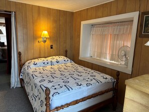 A bedroom with a wooden paneled wall, a bed with a nautical-themed bedspread, a bedside lamp, a window with a lace curtain, and a standing fan. - Stay at "Seas the Day" in Ferry Beach, Saco, ME—an inviting 4-bedroom cottage near Camp Ellis (Saco)