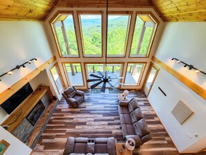 Ein geräumiges Wohnzimmer mit einer großen Holzbalkendecke, einem Kamin, einem Deckenventilator und Blick auf eine bewaldete Landschaft durch große Fenster.