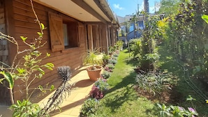A wooden house with a covered porch, a garden with various plants and flowers, and a clear blue sky. - Maison aux Portes du Sud Sauvage – Family Home with Pool & Hot Tub (Saint Joseph)