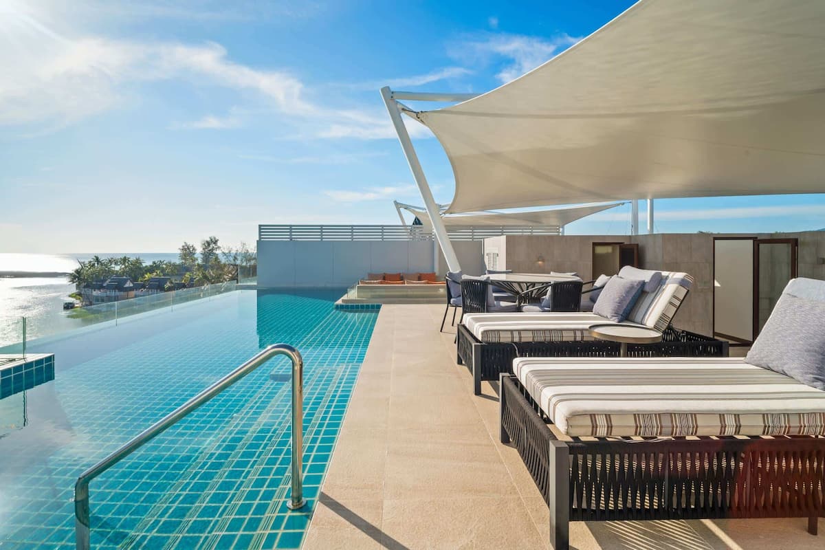 A rooftop infinity pool with a view of the ocean, outdoor seating with cushions, and a large canopy providing shade.