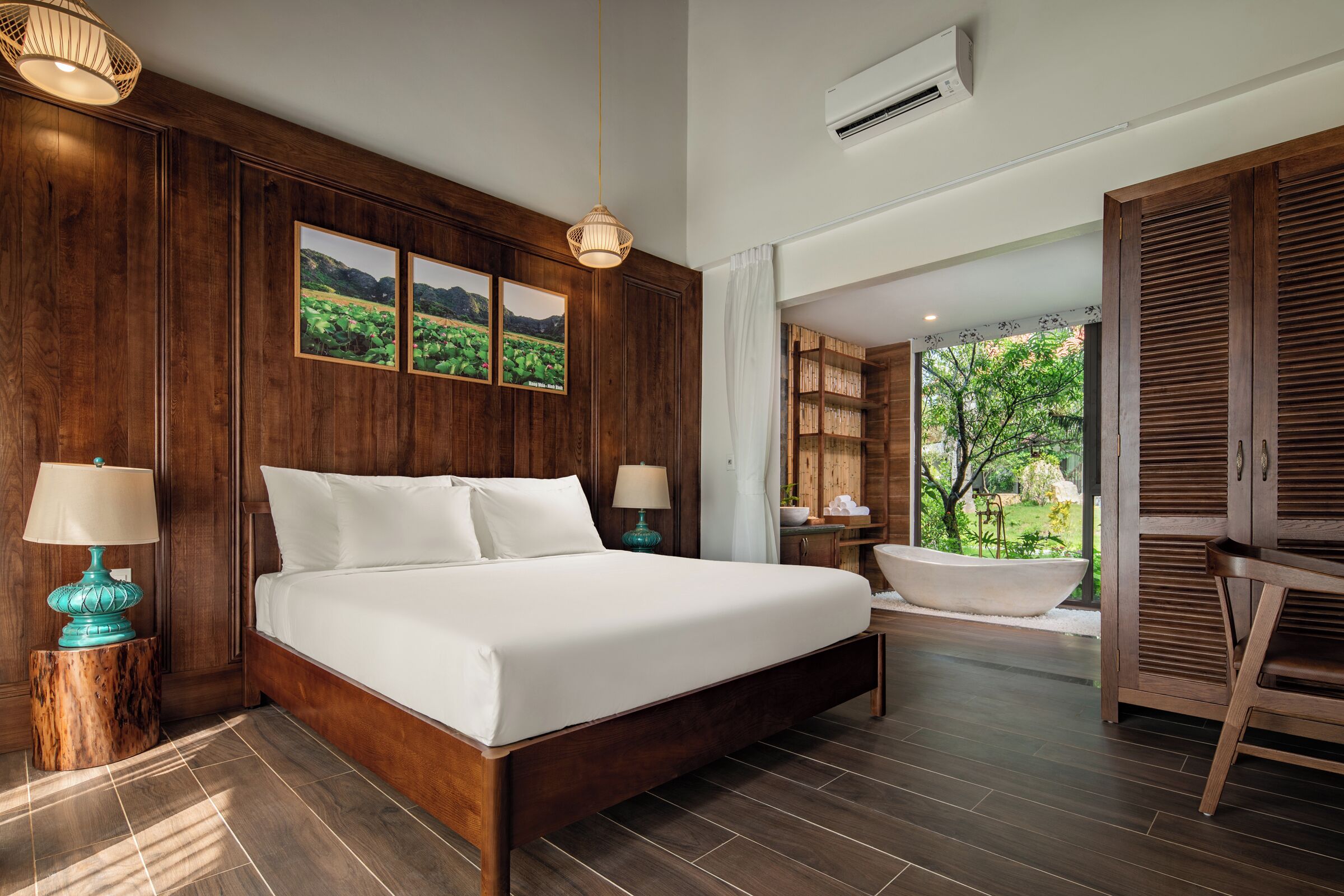 A bedroom with a large bed, wooden paneling, two bedside lamps, a view of a garden through a window, and a bathtub in the corner.