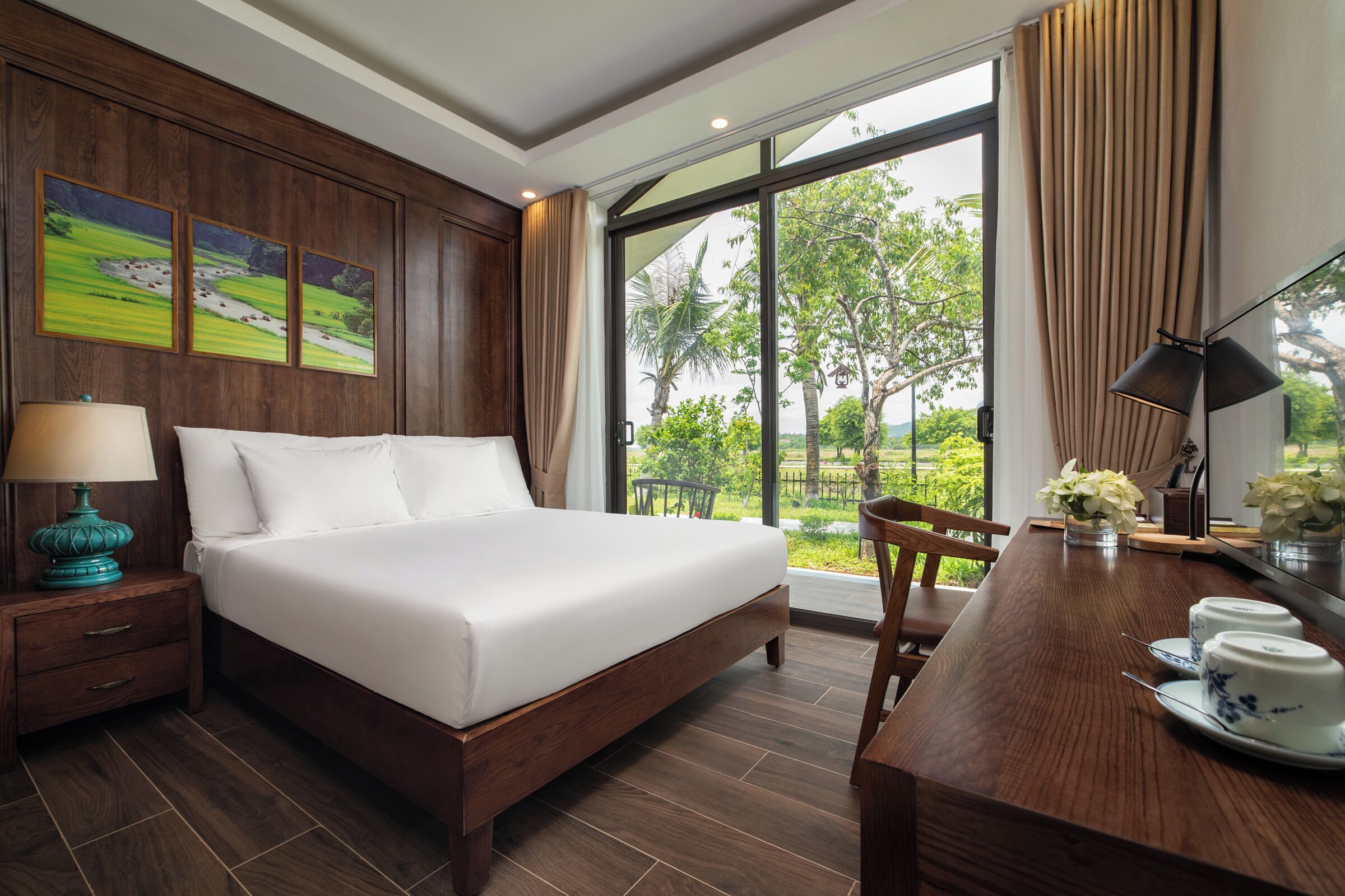 A hotel room with a large bed, wooden nightstand, bedside table with lamp, dining table with chairs, and large windows with a view of greenery.
