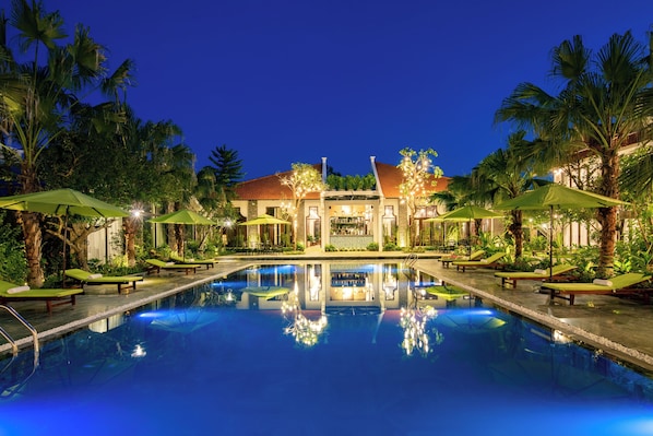 A pool area with lounge chairs and umbrellas, surrounded by palm trees and a building with a red roof.
