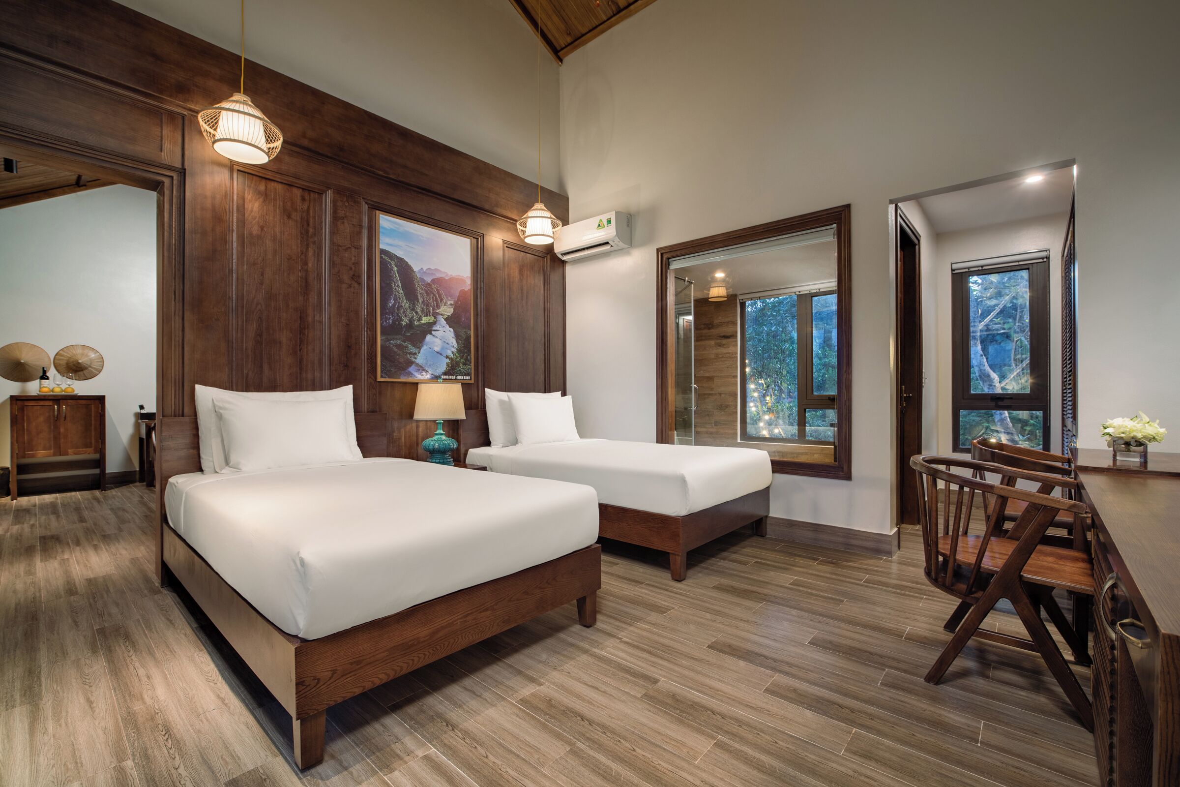A bedroom with two beds, wooden flooring, a dining area with chairs, and large windows.