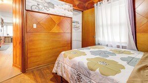 Une chambre avec une armoire en bois, un lit recouvert d’une couvre-lit à motifs floraux et une fenêtre ornée de rideaux légers.