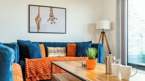 A modern living room with a blue sofa, orange throw, wooden coffee table, and a framed giraffe picture on the wall. - Seascape (Newquay)