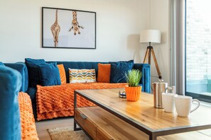 Ein modernes Wohnzimmer mit einem blauen Sofa, einer orangefarbenen Decke, einem hölzernen Couchtisch und einem gerahmten Giraffenbild an der Wand.