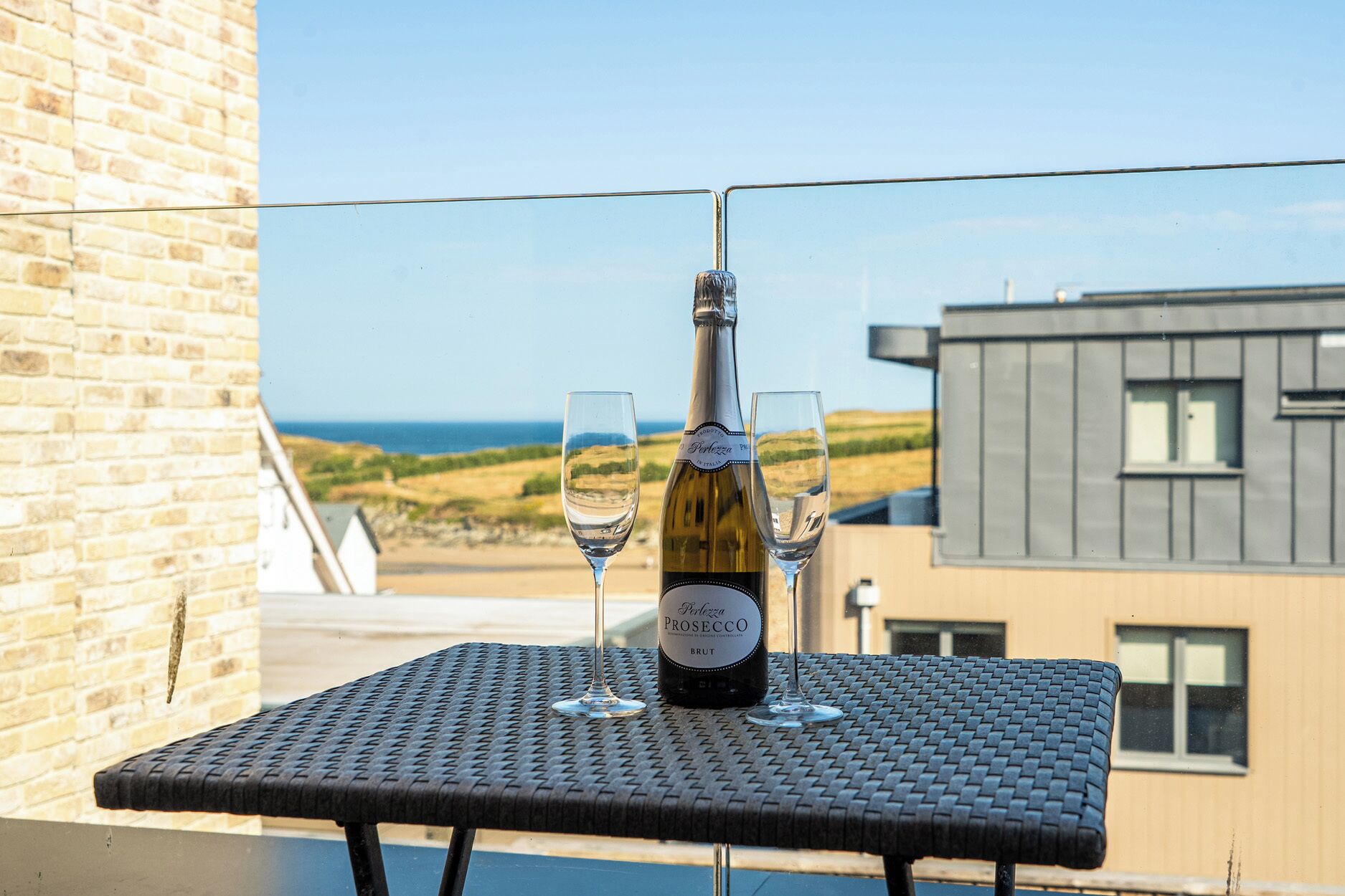 A bottle of Prosecco wine on a table with two glasses, set against a view of the sea and a modern building.