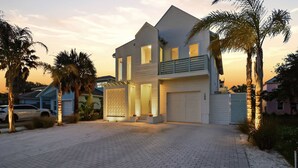 Une maison moderne à deux étages, avec une allée, des palmiers et une voiture garée au premier plan.