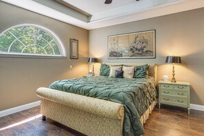 Quarto com cama grande, um banquinho, mesa de cabeceira e janela com vista para árvores.