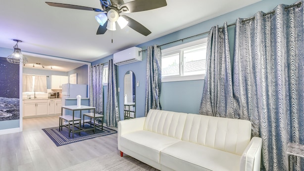Ein Wohnzimmer mit weißem Sofa, Deckenventilator, Esstisch mit Stühlen und einer Küche im Hintergrund.