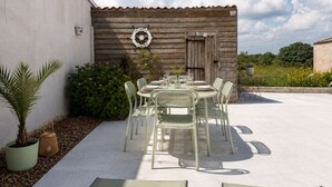 Um pátio externo com uma mesa posta para quatro pessoas, rodeado de cadeiras, em frente a uma parede rústica de madeira com uma placa acima.