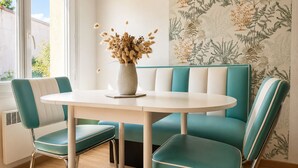 Uma área de jantar com uma mesa redonda, cadeiras azul-turquesa e brancas, e um vaso com flores secas.