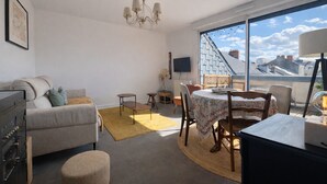 Un salon moderne comprenant un coin repas, un lustre, une grande fenêtre donnant sur la ville et un téléviseur fixé au mur.