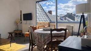Une pièce comprenant une table à manger, des chaises, un téléviseur, un radiateur, une fenêtre donnant sur des immeubles et un lustre.