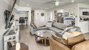 A modern living room with a sectional sofa, a coffee table, a flat-screen TV mounted on the wall, and a ceiling fan.