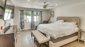 A bedroom with a bed, a bench, a TV, a ceiling fan, and a window with curtains.