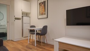 Moderni huone, jossa on ruokapöytä, kaksi tuolia, valkoinen penkki, taulutelevisio, jääkaappi ja seinällä oleva kehystetty taulu.