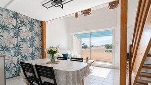 Uma área de refeições com mesa, cadeiras, um vaso de flores e vista para uma varanda através de uma porta de vidro.